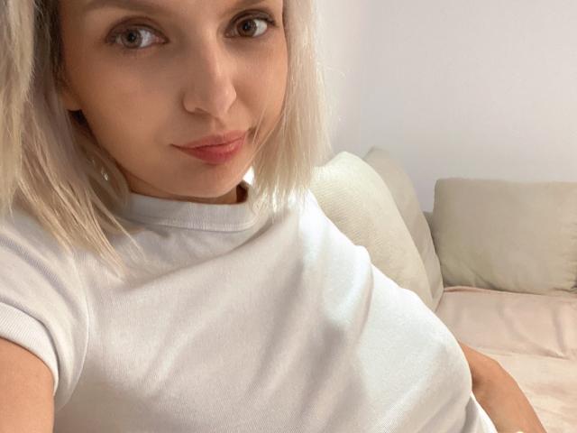 DouceAmelia with blonde hair, wearing a white t-shirt, showing her tits, sitting on a bed, smiling slightly, light brown eyes, fair skin, pink lips, soft lighting.