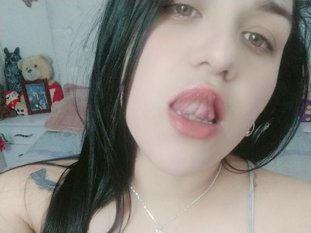 VivianaThompson has pale skin, black hair, and is sticking out her pink tongue with her mouth slightly open. She has a small nose ring and a tattoo on her shoulder.
