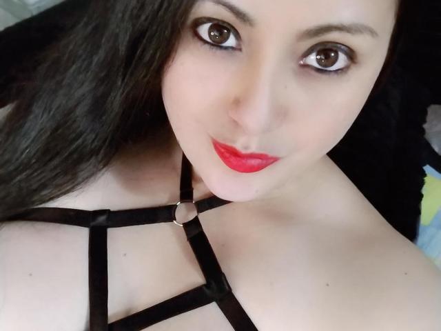 Karol69Sex with dark hair, brown eyes, red lipstick, and black strappy lingerie, showing her bare shoulders and a metal ring on the strap. Her tits are partially visible.