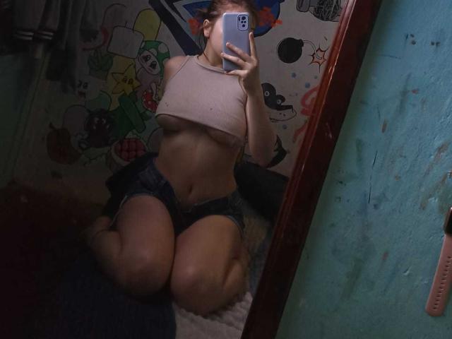 SophieMarkrov knielend voor een spiegel, met een beige topje aan dat haar borsten laat zien, en een denim short, terwijl ze een selfie maakt met een telefoon, waarbij haar gezicht gedeeltelijk verborgen is..