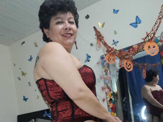 angellovelly, short curly black hair, smiling, red lace-up corset, showing her tits, reflection in mirror, butterflies and Halloween decorations on wall, ceiling tiles, casual indoor setting.