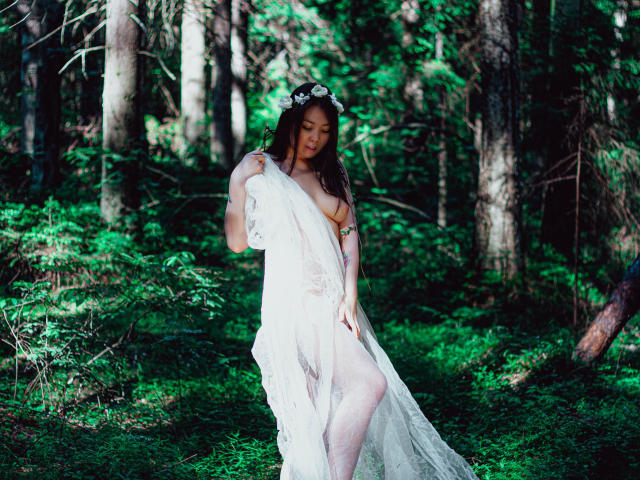 NatzukoShi, con cabello largo y castaño, usando una diadema blanca con flores y un vestido de encaje transparente, que deja ver sus pechos y vulva, está de pie en un bosque verde, sosteniendo su vestido..