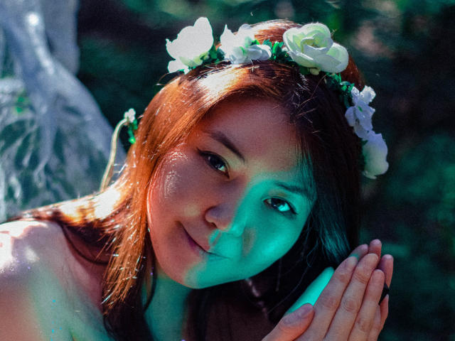 NatzukoShi with brown hair, white flower crown, and green light shadow on face, smiling softly, hands together, sunlight highlighting her skin, lush green background, peaceful expression.