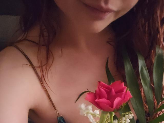 LilouLove's bare shoulder with a pink rose in her hand, holding a blue gem necklace, has wavy red hair, and a subtle smile, showing soft skin and pink lips.