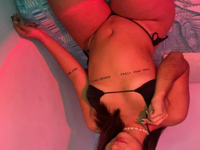 EmmaWaltter lying on her back, wearing a black bikini, exposing her tits and ass, with tattoos on her ribs, holding a green leaf, in red-pink lighting.