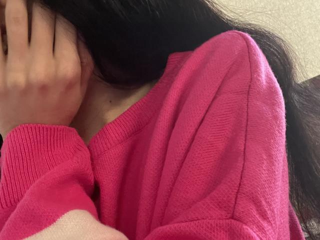 El rostro de RozalinaPleasure se ve parcialmente, vistiendo un suéter rosa, con cabello negro largo y su mano cubriendo su boca. Sus pechos son ligeramente visibles debajo del suéter..