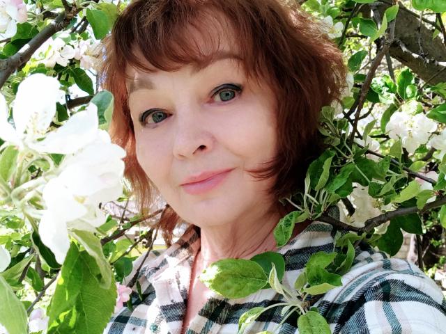 MilaMils heeft kort rood haar, groene ogen en een subtiele glimlach. Ze draagt een groen-wit geruit shirt, omringd door witte bloemen en groene bladeren..