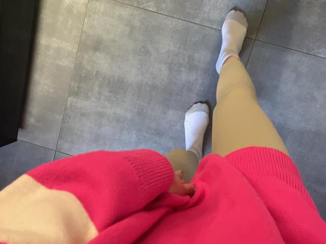 RozalinaPleasure, com as pernas vestidas com leggings bege, meias brancas e um suéter rosa, em pé em um chão de azulejos cinzas, mostrando suas coxas pálidas e uma pequena parte de suas nádegas..