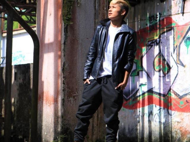 TamarahhRyan stands in a sunlit, graffiti-covered alley, wearing a black leather jacket, white tee, and black pants, with hands in pockets, looking to the side, blonde hair.