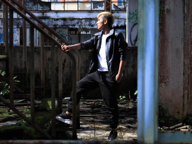 TamarahhRyan with blonde hair, wearing a black jacket, white tee, black pants, and black shoes, stands with one foot on a rusty staircase in a dilapidated, graffiti-covered building.