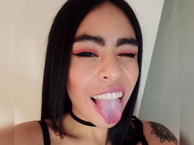 StellaBeckker with black braids, blue eyeshadow, and a tongue piercing, sticking out her tongue and showing a nose ring, on a green couch.