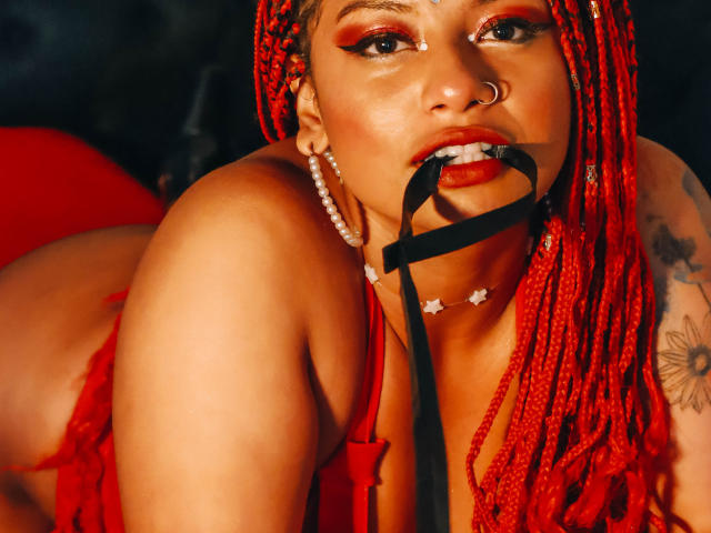 NashaJonny with red braids, red glossy latex, covering her big tits with one hand, wearing matching red wrist cuffs, hooped earrings, and black nail polish, against a warm orange background.