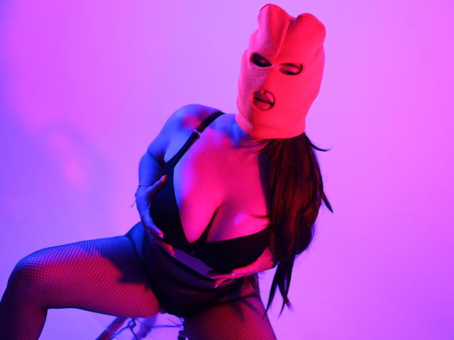 ChristineBray in a pink mask, black bra, and fishnet stockings, with purple and pink lighting, sitting with legs apart, showing her tits and a sexy, seductive expression.