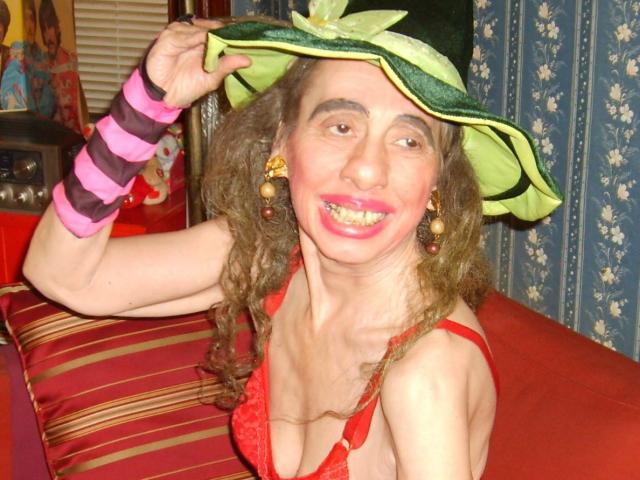 ElenahHottie mit lockigem, braunem Haar, die einen grünen Hut, ein rosa gestreiftes Armband, einen roten Spitzen-BH und pinken Lippenstift trägt, lächelt mit sichtbaren Zähnen und zeigt kleine Brüste..
