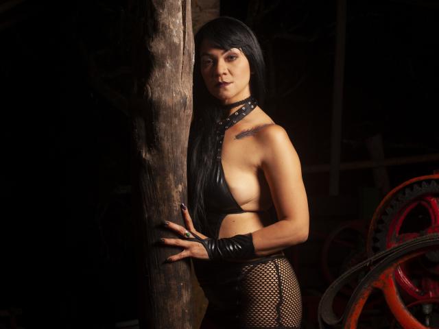 CharlotteJhons, con cabello negro largo, un collar de cuero, una blusa de cuero negro reveladora, pantalones de malla de pescado y guantes negros, de pie contra un poste de madera, mostrando sus pechos pequeños y un tatuaje..