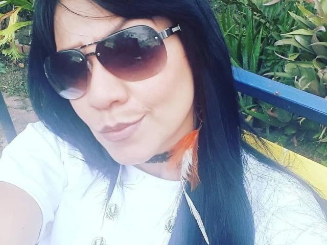 CharlotteJhons, com cabelo preto, usando óculos de sol marrons grandes, camisa branca, colar de penas laranja e uma alça preta, sorrindo levemente. Ela tem um rosto bonito e um visual atraente..