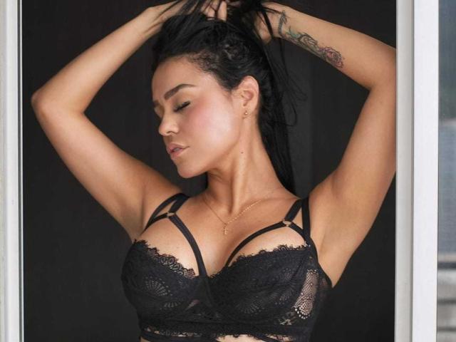 SlinkyAngeel com os braços levantados, mostrando um sutiã de renda preta e seios grandes, pele bronzeada, cabelo escuro em um rabo de cavalo, tatuagem no braço direito, colar de ouro, olhos fechados, maquiagem discreta, confiante..