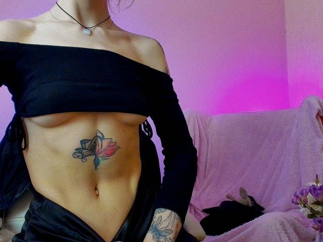 Tsukerberg with off-shoulder black top, exposing small boobs and a tattoo of a pink and blue lotus on her toned, flat stomach. Purple light in the background.