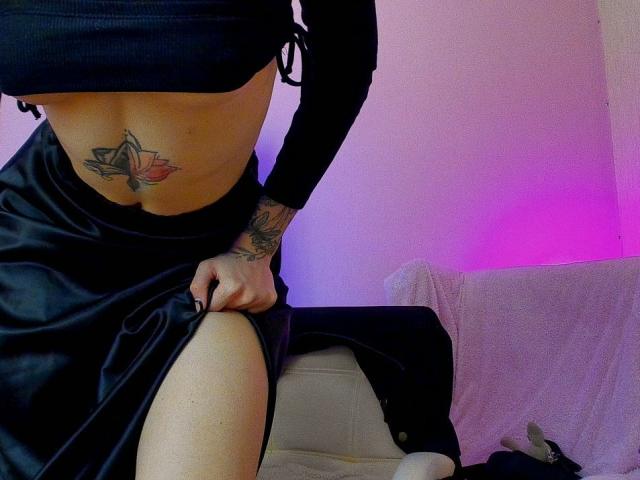 Tsukerberg vestindo uma cropped preta, uma saia preta e com uma tatuagem rosa de uma flor de lótus visível na parte inferior do abdômen, levantando a saia para revelar a coxa. Iluminação roxa..