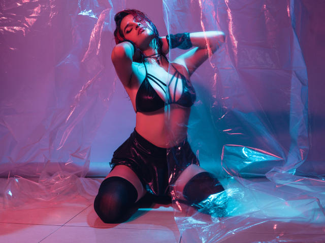 CameroonSweet agenouillée, portant un soutien-gorge noir en cuir à bretelles et une jupe brillante, les mains derrière la tête, enveloppée de plastique brillant, éclairage néon bleu et rose, bas noirs hauts jusqu'à la cuisse, sexy, séduisante..