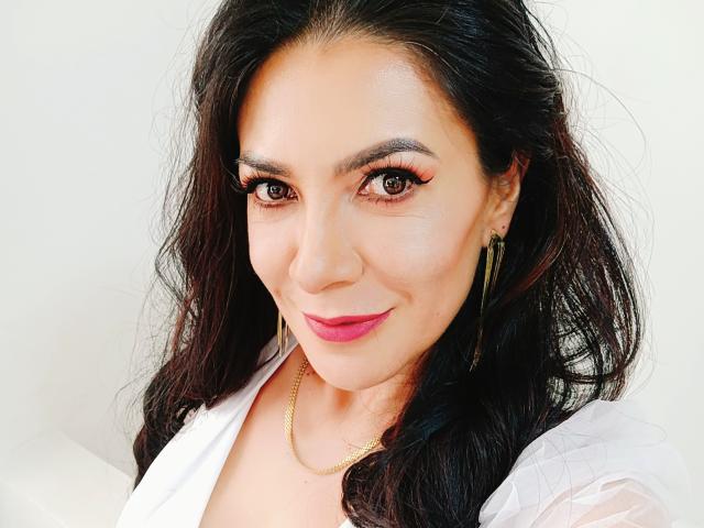 JoTaylor tiene el cabello oscuro y ondulado, ojos marrones con delineador de ojos rosa, lápiz labial rosa, y lleva una camisa blanca con aretes y collar dorados. Está sonriendo suavemente..