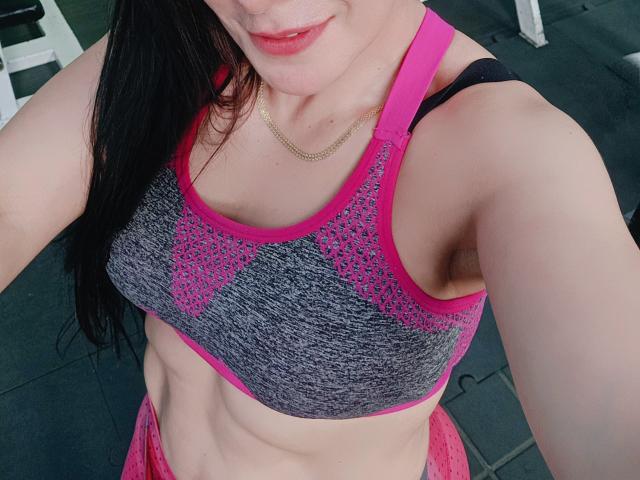 JoTaylor with pale skin, black hair, pink and gray sports bra, showing her boobs and abs, wearing pink workout pants, gold necklace, smiling slightly.