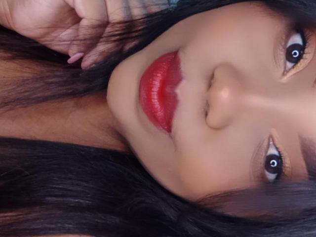 SarahSmity's face with glossy red lips, black hair, and brown eyes, her hand near her cheek, showing pink nails, looking slightly to the side.