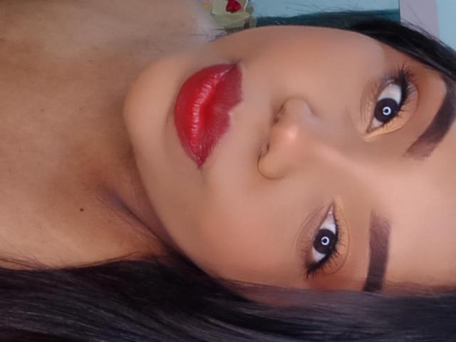 SarahSmity's face, brown eyes, red lipstick, black eyebrows, and dark hair in a selfie, showing her nose slightly to the right, with a soft, smooth skin texture.