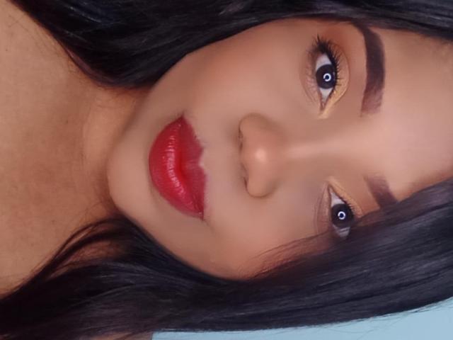 Il viso di SarahSmity, con occhi marrone scuro, rossetto rosso e capelli neri, mostra un naso leggermente all'insù e sopracciglia folte, mentre guarda direttamente la fotocamera..