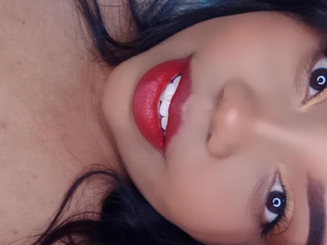 SarahSmity's face, featuring bright red lipstick, white teeth, and dark brown eyes with visible black pupils. She has dark hair and a smooth, tan complexion.