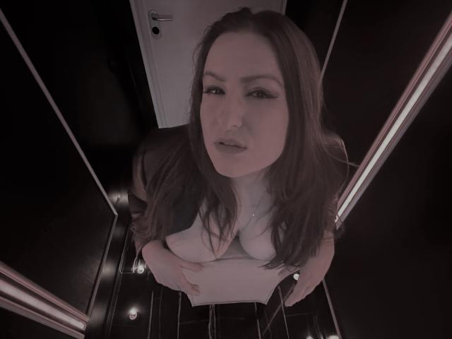 BadAngel with long brown hair, looking up, wearing a white shirt, exposing her tits, standing in a black-tiled, narrow hallway, holding a paper, with a door behind.