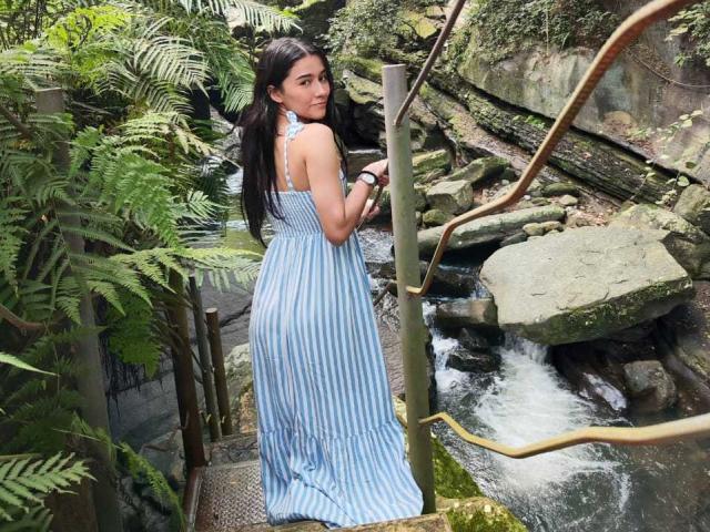 DeisyWest, con cabello negro largo, vestida con un vestido azul a rayas, mirando por encima del hombro, de pie en una escalera de metal junto a un arroyo rocoso con vegetación..