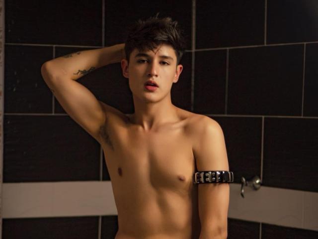 AngelCossio, shirtless with a black leather cuff on his right arm, has short brown hair, a tattoo on his left arm, and a serious expression, standing in a dimly lit room.
