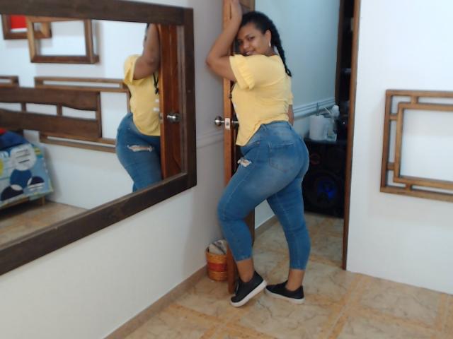 CataleyaX69 debout, dos à la caméra, chemise jaune, jean bleu déchiré, baskets noires, regardant par-dessus son épaule, mains sur la porte, reflet dans le miroir montrant ses fesses..