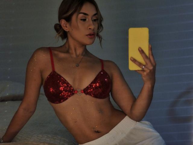 AliceSchmit, avec un soutien-gorge rouge pailleté, tenant un téléphone jaune, pantalon blanc, peau claire, cheveux bruns attachés en chignon, petit piercing au nombril, sexy, regardant le miroir, pièce faiblement éclairée..