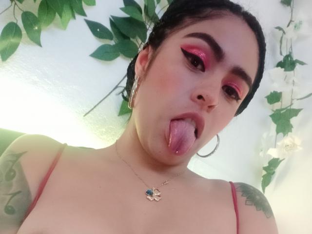 StellaBeckker with pink eye makeup, tongue out, small nose piercing, hoop earrings, necklace, tattoos on arms, bra straps visible, green leafy decorations in background.