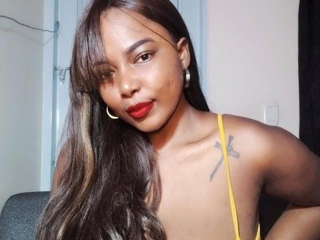Emira, com cabelos castanhos longos, batom vermelho, brincos de argola, blusa amarela, seios pequenos, tatuagem no ombro direito, sentada, olhando para a câmera, sorrindo..
