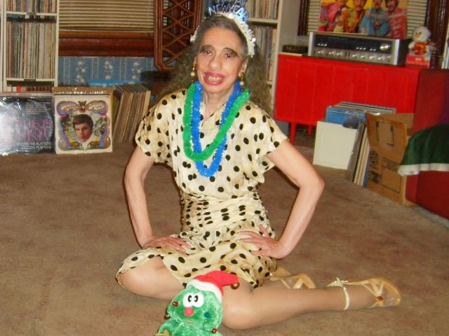 ElenahHottie sitting on carpet, wearing a polka dot dress, green and blue boas, white flower headpiece, tan heels, hands on knees, with green hat on her right thigh.