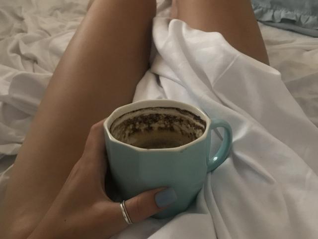 AurikaBriz's hand holds a blue coffee cup with brown crema, resting on white sheets, her bare, tan legs stretched out. She's wearing a silver ring on her index finger.