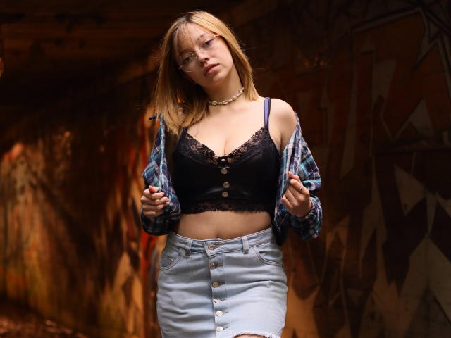 SophieMarkrov, com cabelo loiro, óculos, sutiã de renda preta, camisa abotoada e jeans azuis, expondo seus seios, usando um colar, em uma área urbana, coberta de grafites, com pouca luz..