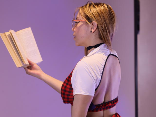 SophieMarkrov with blonde hair, glasses, and red plaid bra, reading a book, wearing a white top with black straps, showing her back and small tits.