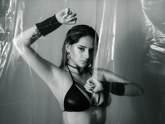 Foto em preto e branco de CameroonSweet com cabelo molhado, usando um sutiã de couro preto, pulseiras e brincos, segurando plástico, mostrando tatuagens e uma expressão séria, com seios visíveis..