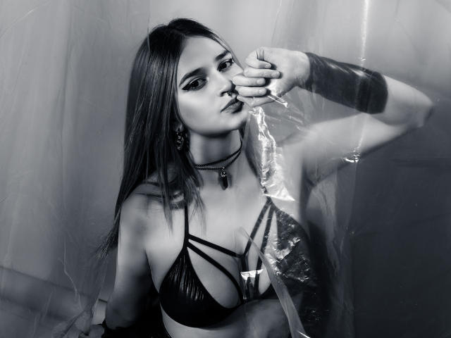 Fotografía en blanco y negro de CameroonSweet con cabello largo, vistiendo un sostén negro con tirantes, un collar y una pulsera, mordiendo una lámina de plástico transparente, mostrando sus pechos, con una expresión seria..