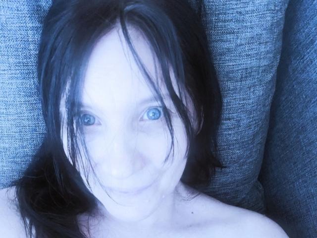 RinaBellas mit blauen Augen, schwarzen Haaren und einem leichten Lächeln, liegt auf einem grauen, strukturierten Sofa, oben ohne, wobei ihre nackten Schultern zu sehen sind..