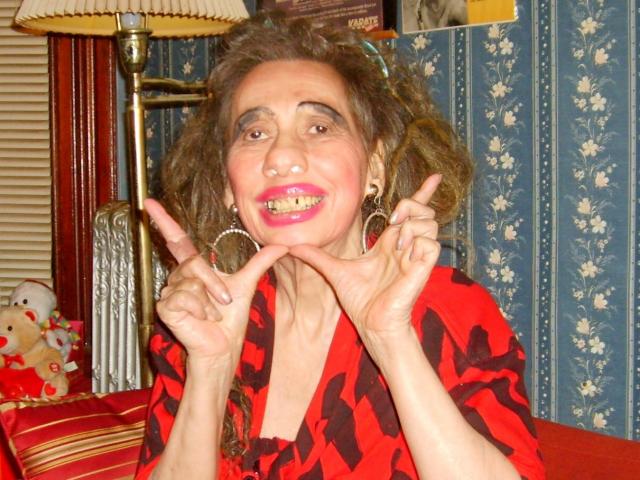 ElenahHottie has curly brown hair, red lipstick, and is wearing a red and black patterned top, making a goofy face with her fingers pointing at her lips.