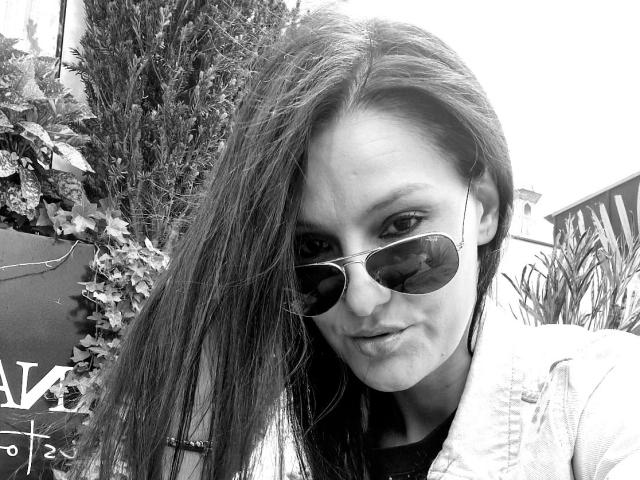 Black-and-white selfie of JoliDanielle with long hair, wearing aviator sunglasses and a denim jacket, showing a slight pout, with a subtle smirk on her face.