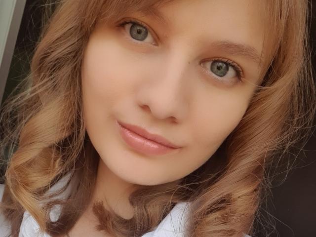 LinNatty's face with light brown, wavy hair, green eyes, and pink lips. Her smooth, fair skin and subtle smile are prominent. She's wearing a white shirt.