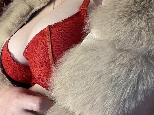 Der rote Spitzen-BH von YourTalisman, der ihre großen Brüste zeigt, unter einem flauschigen, beige gefärbten Fellmantel. Ihre Hand liegt auf ihrer Hüfte, und sie trägt eine zarte goldene Halskette..