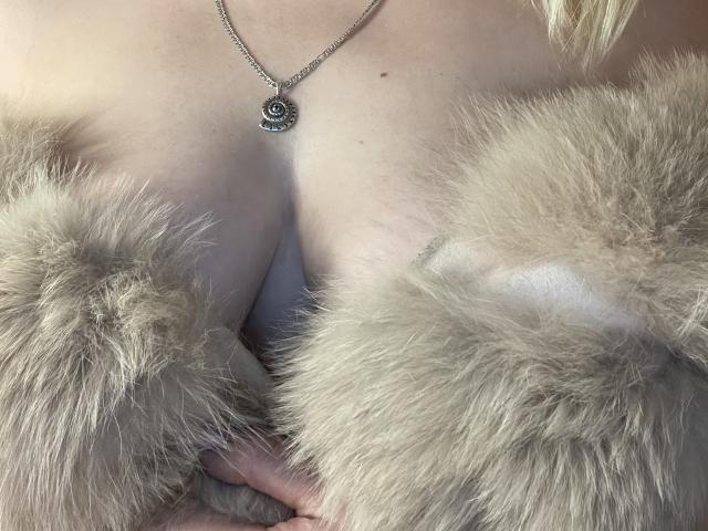 Die Brüste von YourTalisman, bedeckt mit flauschigem, grauem Fell, mit einer silbernen Halskette mit einem schwarzen Rosenanhänger, sichtbare Dekolleté und blonden Haaren, die leicht im Bild zu sehen sind..
