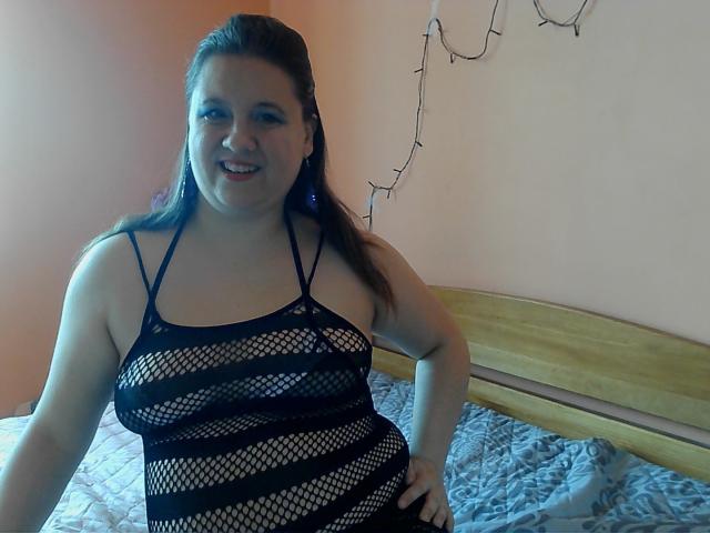 WendyStacy, a plus-sized woman with long brown hair, wears a black fishnet dress showing her big tits and a smile, standing in a bedroom with a wooden bed.